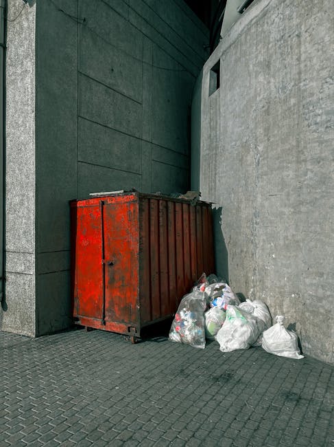 A red metal dumpster with visible rust and chipped paint is positioned against a dark, textured wall made of large, rectangular stone panels on the left side of the image, and a smooth, light grey concrete wall on the right. Surrounding the base of the dumpster are several tied black and white plastic rubbish bags filled with waste, some of which are leaning against the container. The ground consists of dark paving stones arranged in a grid pattern, with a slight incline leading to a narrow alleyway or corner. In the background, part of a small, square window or ventilation opening can be seen high up on the grey wall. The setting appears to be an urban back alley or service area, where waste from local properties is temporarily stored before collection, reflecting a typical environment managed by private waste removal services such as Waste Removal Barnet, serving the rubbish disposal needs of the area. The scene is illuminated by natural daylight, highlighting the textures and weathered condition of the overall scene.