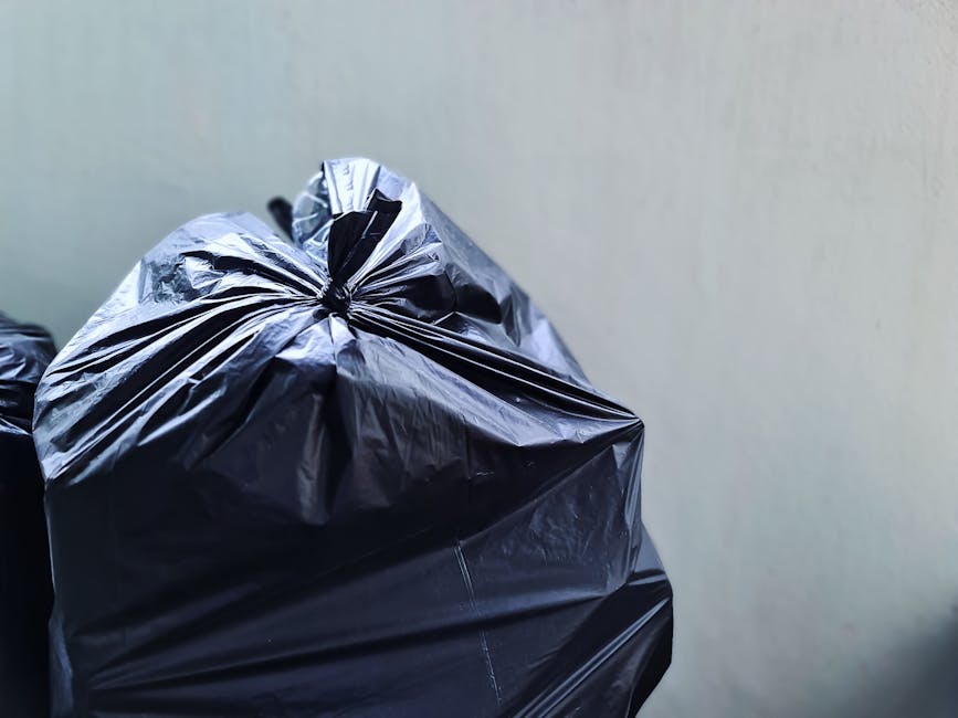 Close-up view of a large black plastic rubbish bag filled with waste material, tightly knotted at the top to secure its contents, positioned against a plain, light-colored wall. The bag has a glossy, smooth surface with visible creases and reflective highlights, indicating it is made of durable plastic. It is situated outdoors on a driveway or pavement area, with other partially visible black bags nearby, suggesting a collection for rubbish removal services. The background includes a neutral, unobtrusive backdrop, emphasizing the bag’s prominence. The scene reflects an instance of on-site waste clearing, common in private waste disposal or alternative rubbish collection efforts by companies like Waste Removal Barnet, which facilitate non-local authority waste handling. The environment appears well-lit with natural light, underlining the practical and straightforward nature of rubbish removal tasks.