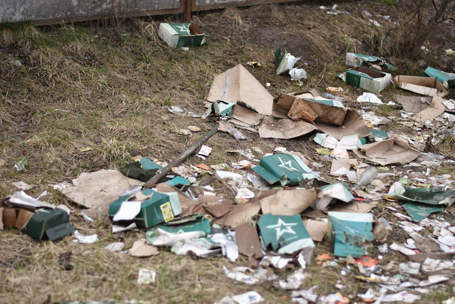 An outdoor scene displaying scattered cardboard boxes, some flattened and torn, alongside crumpled paper, plastic packaging, and debris spread across a patch of grass and soil. The cardboard boxes are predominantly green with white star logos, with some showing signs of weathering and damage. The debris includes torn paper, plastic bottles, and fragments of packaging material, strewn haphazardly on the ground, indicating recent or unmanaged waste. In the background, there is a wooden or metal fence, and the environment appears to be an unmanaged area or roadside verge. The lighting suggests overcast conditions, with diffused natural light highlighting the clutter. This image illustrates the type of unsightly rubbish that may result from illegal dumping or inadequate waste disposal, linking visually to private or alternative rubbish removal services such as those offered by Waste Removal Barnet, who handle bulky waste clearance and junk disposal efficiently outside regular waste collection channels.