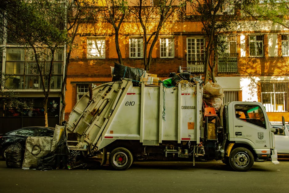 A small white waste collection truck is parked along a street, with its rear compartment filled with various types of rubbish, including black garbage bags, cardboard boxes, and loose waste items. The truck's side panel displays identification marks and the brand name 'Doreabal,' indicating it is part of an independent waste removal service. The waste is piled high above the truck's open container, spilling slightly onto the street pavement. To the left of the truck, a black compact car is parked, with a deck of garbage bags leaning against it, partly obscured. The background features a residential brick building with a warm, orange-brown facade, multiple windows with white frames, some with curtains or blinds, and small balconies with metal railings. Several trees with green foliage cast dappled shadows over the scene, suggesting late afternoon sunlight. The image conveys an scene of private rubbish collection, highlighting the on-site disposal process handled by Waste Removal Barnet in the local area.