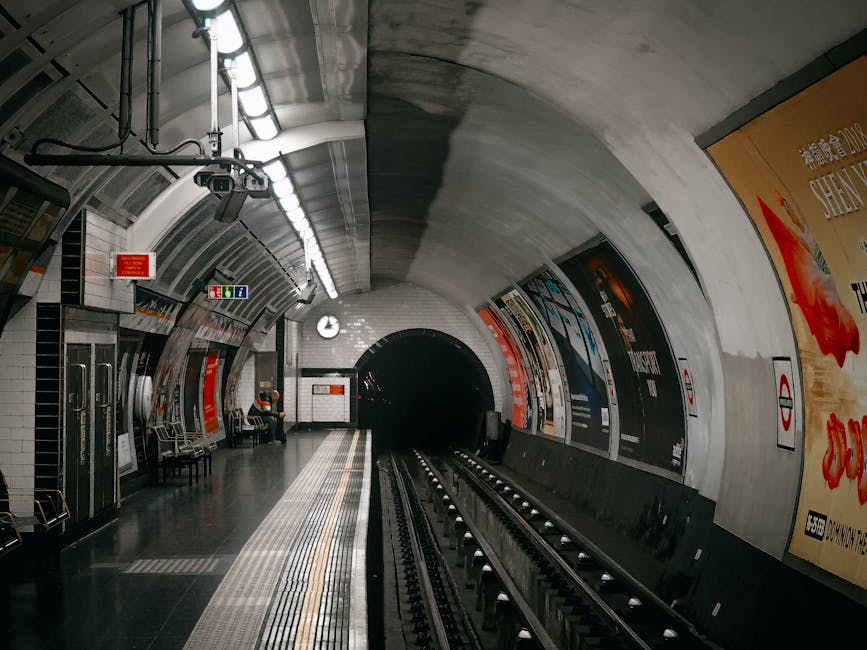 A view of an underground train station platform with a curved, white ceiling featuring industrial lighting fixtures and ventilation systems. On the left side, there are safety signs, seating arrangements, and a small group of passengers, one of whom appears to be waiting or reading. The platform's edge is marked with tactile paving for safety, and the tracks extend into a dark tunnel in the background. The station wall on the right displays large, colorful advertisement posters, and the overall atmosphere is clean and well-lit. This setting exemplifies an urban transit environment typically involved in public transportation, which may connect to the broader context of transport infrastructure often associated with various waste management or disposal services, including those by Waste Removal Barnet for local or private waste collection needs near transport hubs.