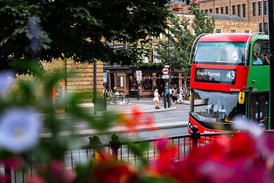 A double-decker bus, predominantly red with green accents, is visible on a busy urban street near High Barnet tube station, with the destination display reading 'Friern Barnet 43'. The bus is positioned on the right side of the image, partially obscured by a black metal fence and out-of-focus colorful flowers in the foreground. In the background, there are pedestrians crossing the street, some standing and others walking, along with bicycles parked near a building with a wooden facade. The environment features a mix of brick and stone architecture, with several shopfronts and signs, indicative of a lively commercial area. Trees with green foliage provide partial shade, and the ambient lighting suggests daylight with a slightly overcast sky, typical of a city centre setting. This scene aligns with the context of everyday urban transportation and local waste management considerations, as indicated by the presence of Waste Removal Barnet’s services in a busy, well-maintained street scene.