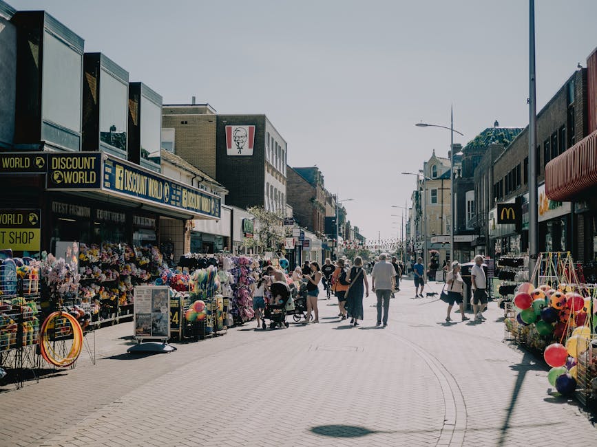 A busy street scene on Barnet High Street showing various retail shops and vendors. In the foreground, there are market stalls on both sides of the paved pedestrian walkway, displaying an assortment of colorful household items, toys, and accessories, with some items stacked or hanging from display racks. To the left, a stall features nylon hula hoops and plastic balls, while further along, there are vendors selling artificial flowers and small decorative objects. On the right side, additional market stands display balloons and other merchandise. Behind the stalls, there are multiple storefronts, including a discount shop with blue signage, a fast-food outlet, and a McDonald's restaurant. The surroundings include mid-rise brick and concrete buildings with large glass windows, some featuring signage and advertisements. Several pedestrians are walking along the street, some pushing strollers or carrying shopping bags, creating a lively urban atmosphere on a clear, bright day. The scene captures a typical high street environment, where independent collection or private waste handling might be required for discarding packaging or unsold goods, aligning with rubbish removal services.