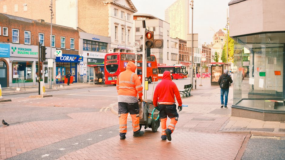 Two waste removal workers dressed in high-visibility orange and red jackets, with matching reflective trousers, are walking away from the camera along a city street, pushing a trolley loaded with trash or recycling bins. One worker is holding the handle of the trolley, while the other appears to be guiding it. The background features a busy urban environment with red double-decker buses, commercial storefronts including a building society and a shop named Coral, and pedestrians walking on the pavement. There is a traffic light with red signals overhead, and various shop windows and street furniture such as benches. The scene is illuminated by natural daylight, with a mix of modern and older architectural buildings along the street, providing a typical setting for private waste collection or rubbish removal services in a metropolitan area, exemplifying an independent approach to urban rubbish disposal within a busy city environment.